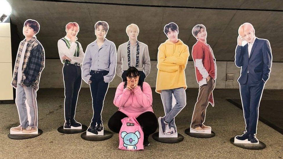 Mel is crouched down on the floor and is posing in front of 7 cardboard cut outs of the South Korean Boyband BTS. She is wearing a pink hoody and black jeans.