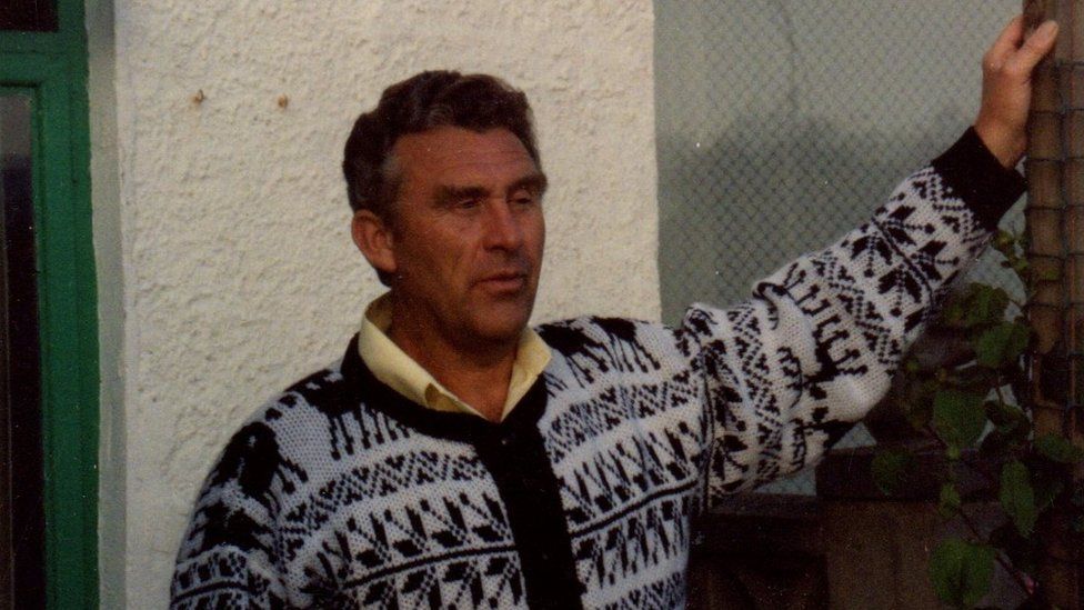Robert Murray pictured in 1992. He is standing outside a house wearing a black and white Christmas jumper with reindeer on. He is holding onto bamboo fencing with one arm and looking into the distance