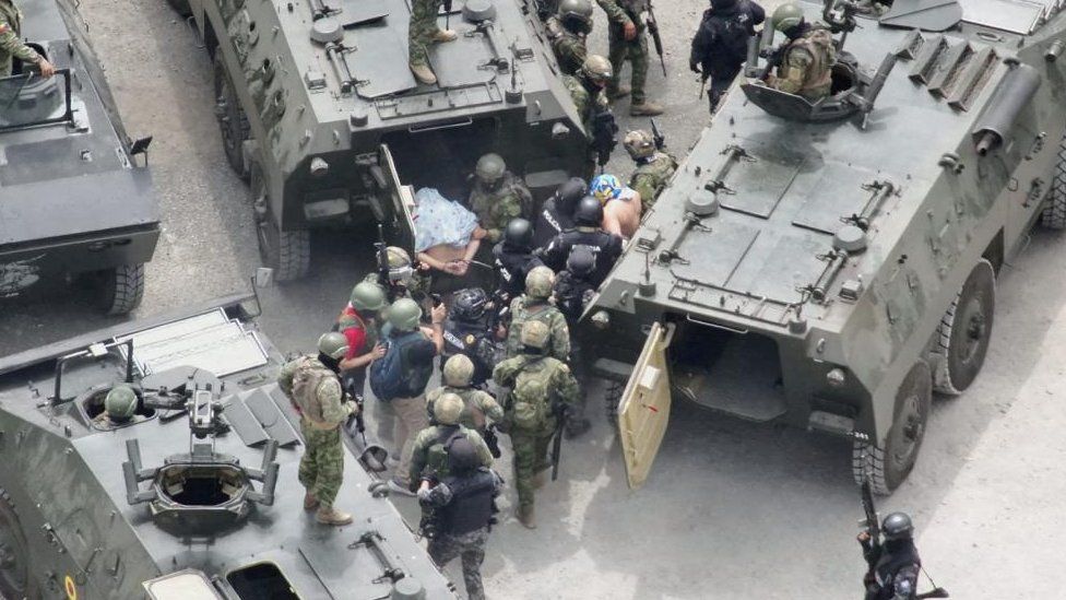 Two handcuffed men with shirts over their heads are moved into tanks by Ecuador police