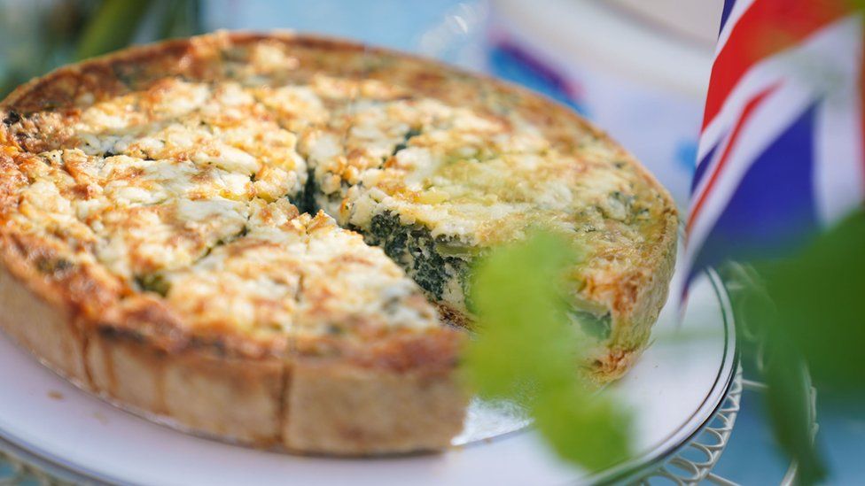 A Coronation Quiche missing a piece sits on a table, next to a UK flag