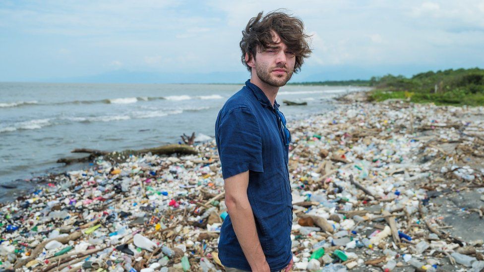 Boyan Slat stands on a beach covered in plastic bottles and items in Honduras