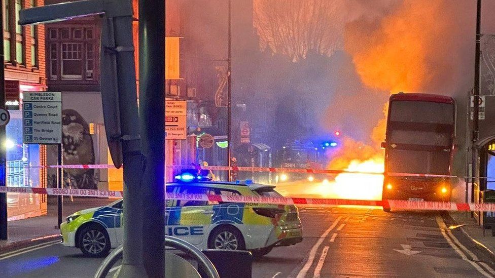 View of a street in Wimbledon filled with smoke and the glow of flames behind a bus
