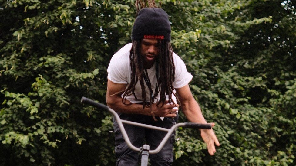 Andre Gillies, a black man in his early 20s, is pictured riding hands free on his BMX bike. Andre wears a black beanie hat with 'Chicago' stitched in red on the front. His hat covers his long dread locks down to his middle. He also wears a plain white t-shirt and black jeans. He is looking down at his handle bars and has a focussed expression with his mouth slightly open. He is photographed outside in front of a green hedge which fills the backdrop