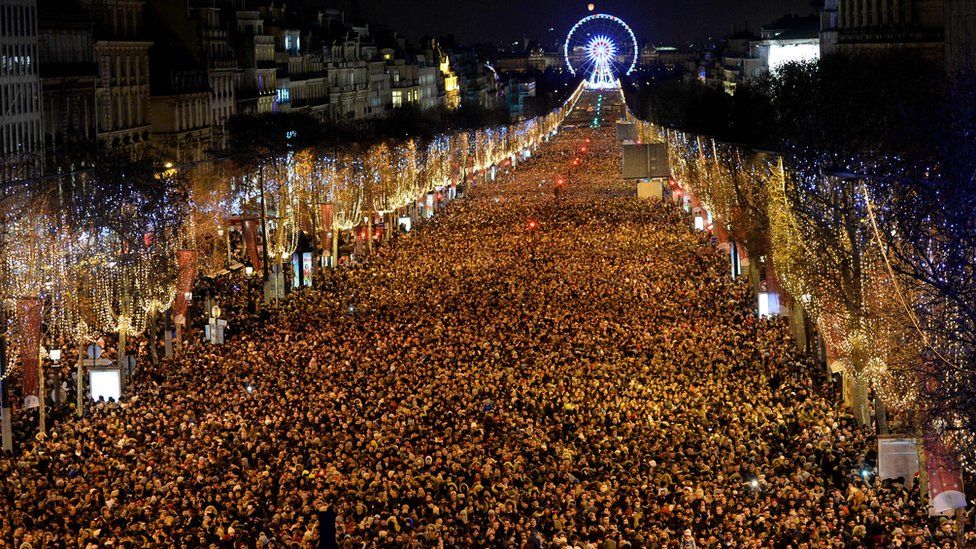 Thousands of people stand on the long avenue in central Paris as far as the ye can see, with a distant Ferris wheel in the background
