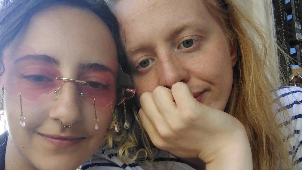 A selfie of couple Daisy, 24 and Ramona, 25. Daisy has her dark hair pulled up, and is wearing pink tinted sunglasses with jewels dangling underneath them and has a nose ring piercing. Ramona has long, straight blonde hair, and is wearing a white T-shirt with horizontal navy stripes. She also has her hand resting on her chin blocking her mouth.