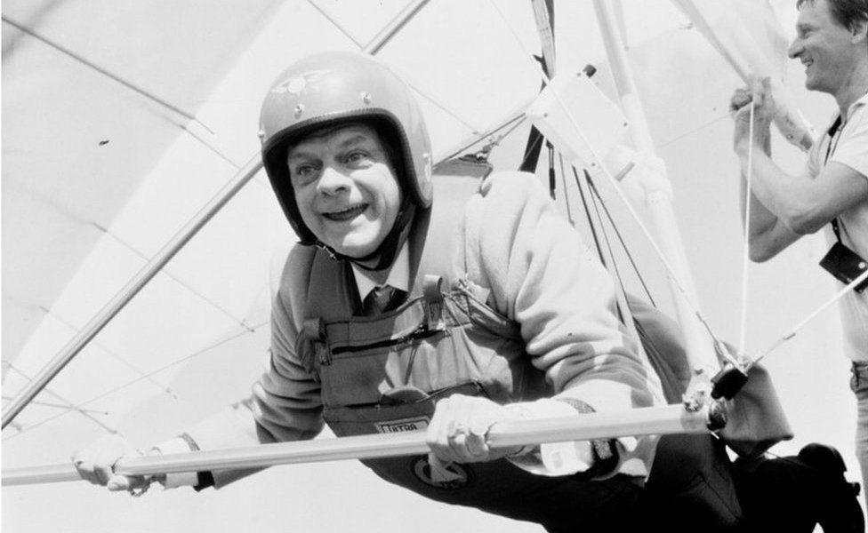 Black and white photo of David Jason holding a hang-glider while being assisted by a member of the production team