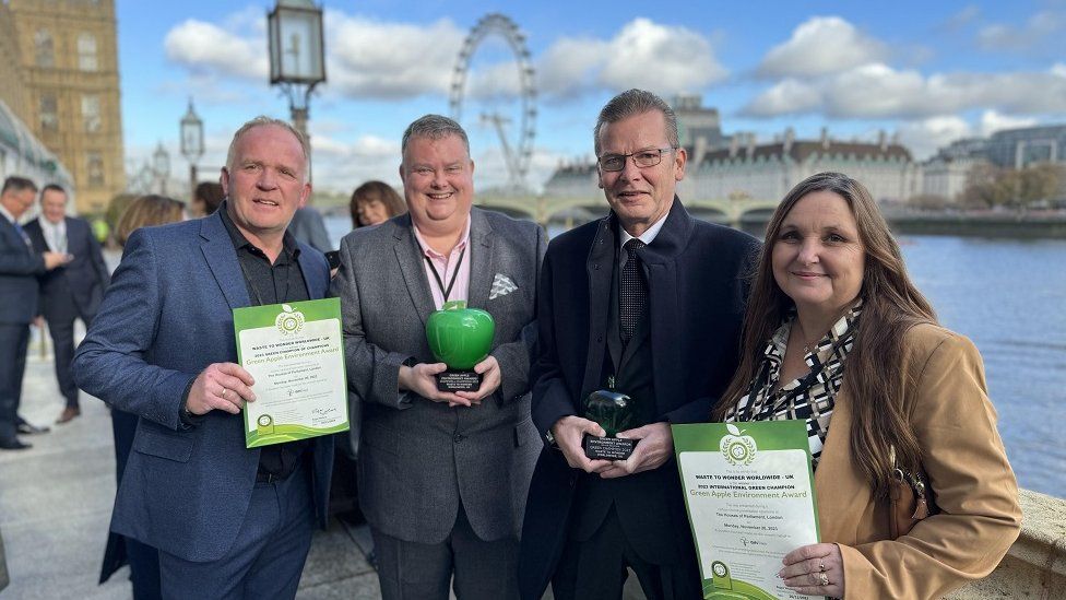 Four people standing with a view of London behind them, including the London Eye, holding green apple ornaments and certificates
