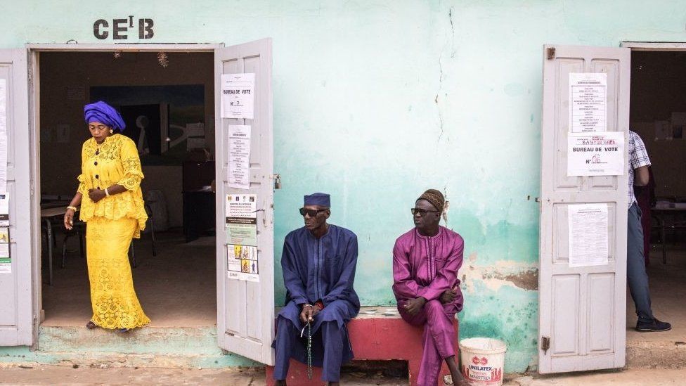 Two men sitting outside a polling booth, they are sitting down wearing traditional West African clothes. There is a woman walking out of the building to the left. She is wearing a yellow outfit with a blue head dress.