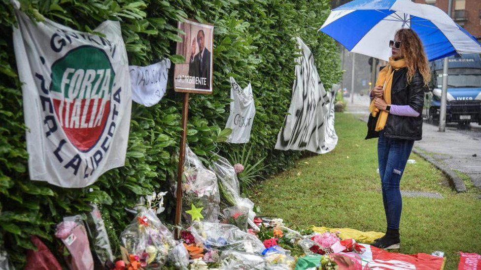 A woman holding an umbrella stands in front of tributes and flowers placed near the entrance to Villa San Martino in Arcore, where the coffin of Silvio Berlusconi was brought earlier in the day,