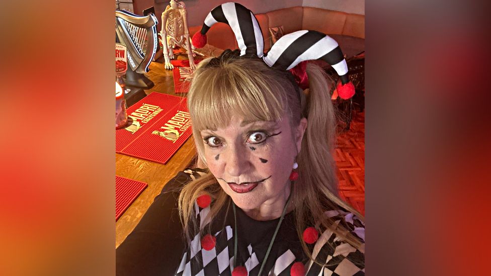 A photo of the landlady in a black and white jester costume in front of the bar