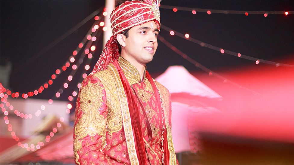 A male groom dressed in South Asian outfit. There is a sparkly red headwear with golden patterns running across. He is wearing a golden and red outfit with intricate and shiny patterns running throughout. Behind him are red lights decorating the wedding venue.