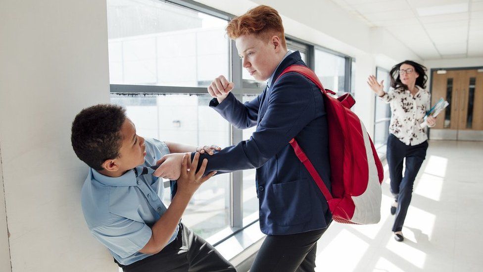 Boy hitting another boy with a teacher running down the corridor to intervene