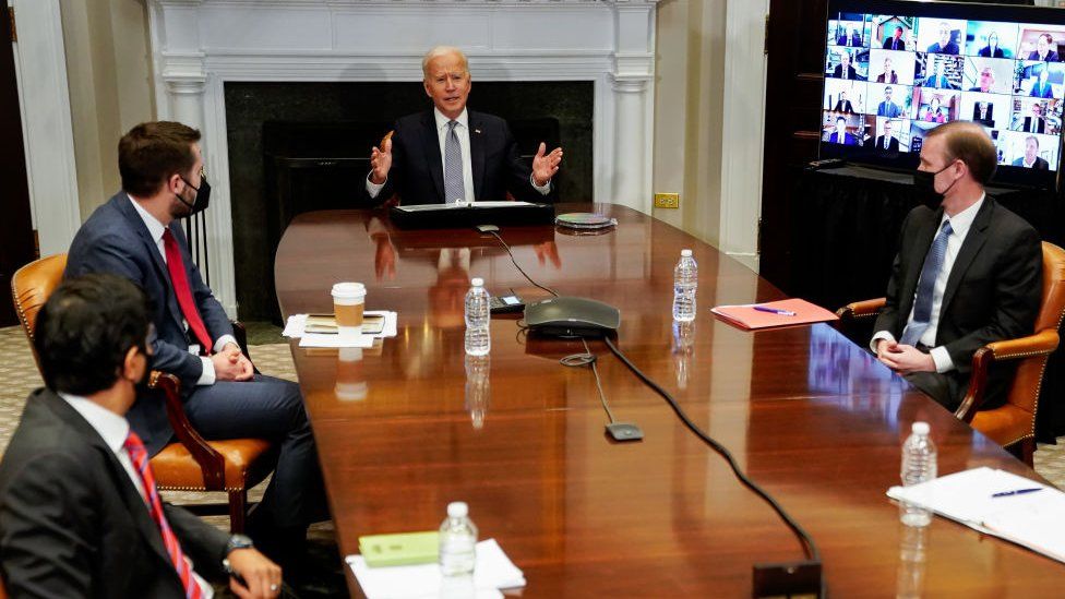 President Biden and three advisers sit around a table as business leaders join by video link