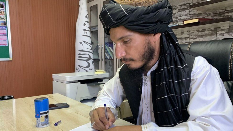 Айнудин сидит за деревянным столом с черно-белым флагом талибов за спиной