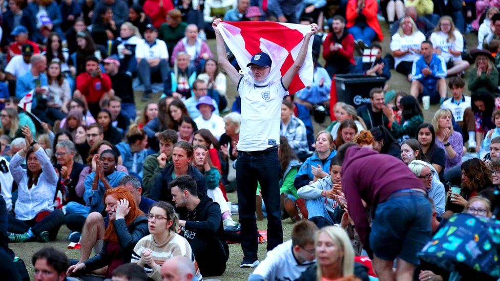 An England fan standing in a crowd holds an England flag while watching the team play Sweden in the women's Euro 2022 semi-final