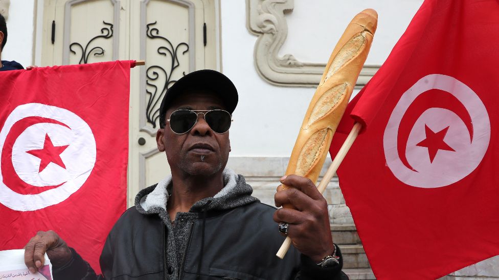 A man commemorating the 1986/1984 bread riots holds up a baguette in front of the Tunisian flags in Tunis, Tunisia - Monday 3 January 2022