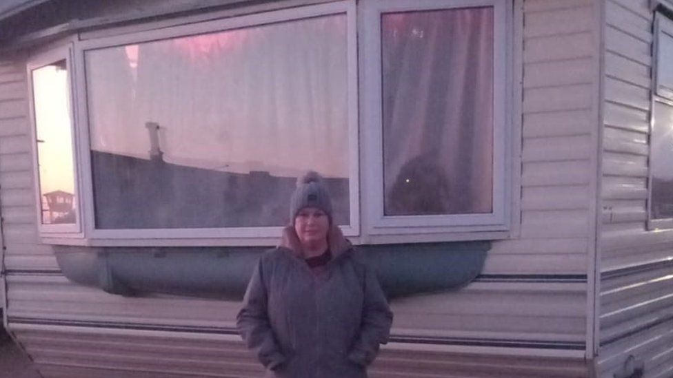 Nichola Wilson, wearing a bobble hat, stands outside the window of her caravan