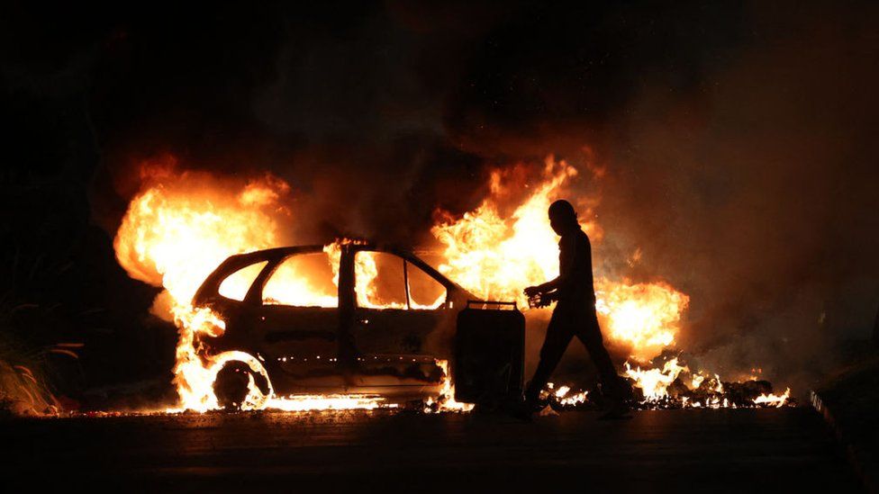 A protester walks by a burning car during clashes with police in Le Port, French Indian Ocean island of La Reunion, on June 30, 2023, three days after a 17-year-old boy was shot in the chest by police at point-blank range in Nanterre, a western suburb of Paris.