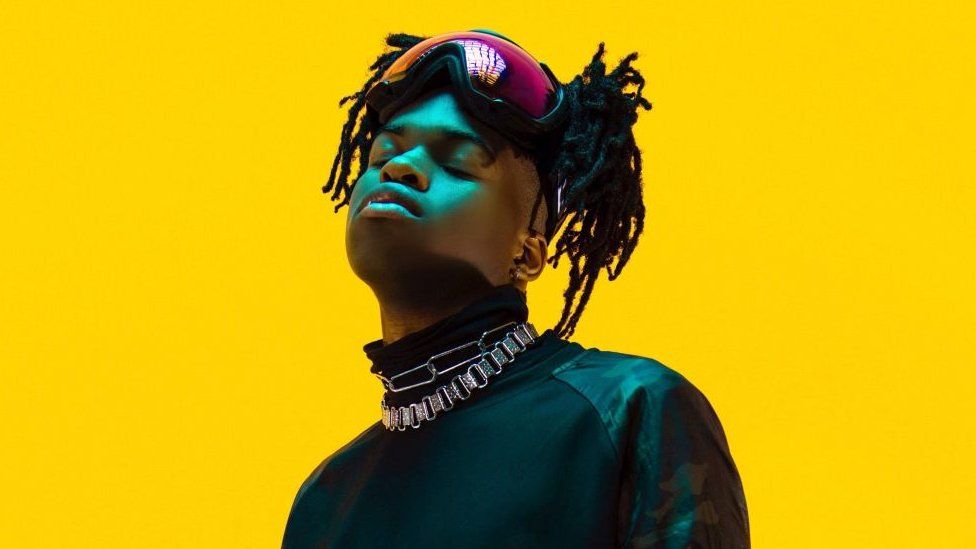 Jordan Adetunji, a black man in his 20s, poses wit his eyes closed against a bright yellow backdrop. Jordan, who has his braided hair tied up, wears ski goggles on top of his head, a high necked black top and two chunky silver chains around his neck. He looks up, slightly angled to the left of the camera, with his face lit by a blue/ green light