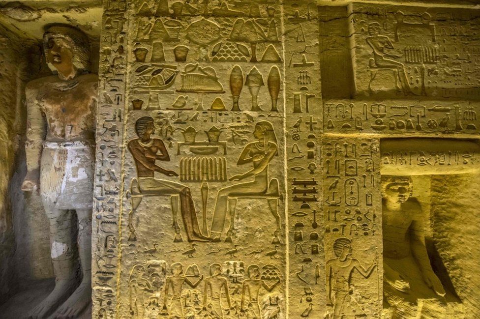 A wall covered in engraved hieroglyphs, including people, weighing scales, birds and what appears to be food