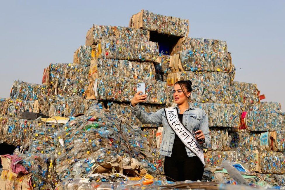 Miss Egypt Hager Mohamed wearing a jean jacket and black dress taking a photo in front of a mountain of plastic rubbish. She is wearing a sash which reads: Miss Egypt 2022.