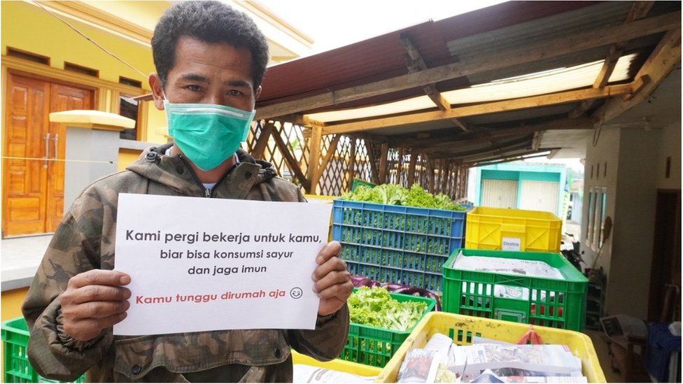 Farmer Pak Opik holds up a sign that says "We go to work for you, so you can have vegetables to stay healthy"