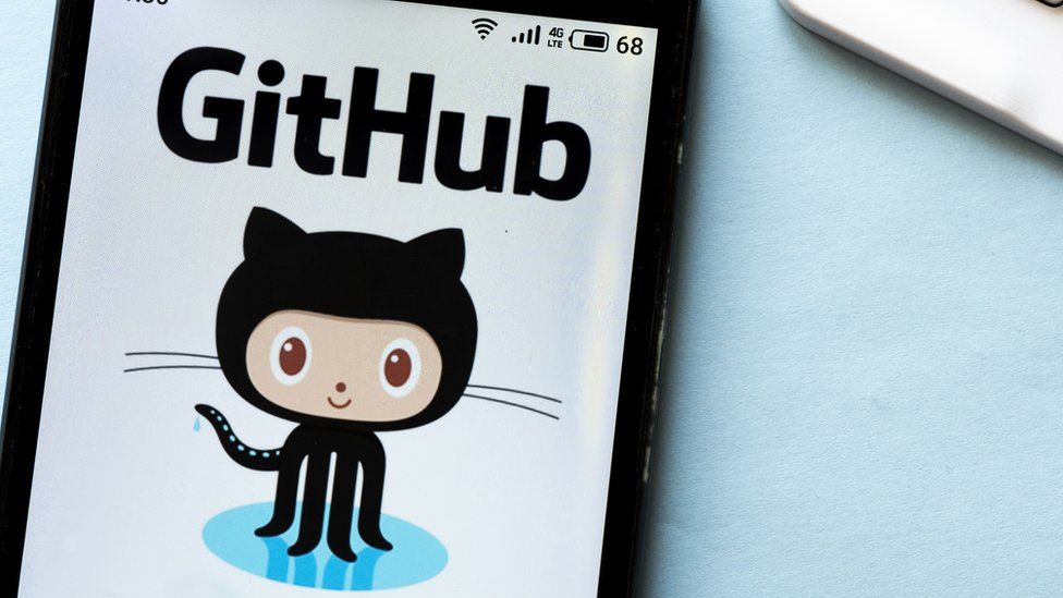 the Github logo is pictured here - a hybrid octopus / cat creature with a nearly-human face, in cartoon style. It is seen on a phone against a light blue table near a laptop