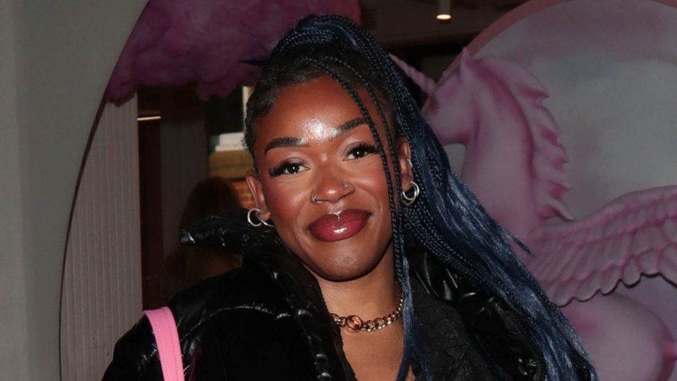 Jasmine Boatswain pictured after The Traitors. Jasmine is a black woman in her 20s with long braided hair. She has brown eyes and smiles at the camera, her head tilted to the left. She wears hooped earrings and also has her nose pierced. She wears a pink bag over a black jacket. She's pictured outside at night, behind her a window display features an image of a pink Pegasus unicorn.