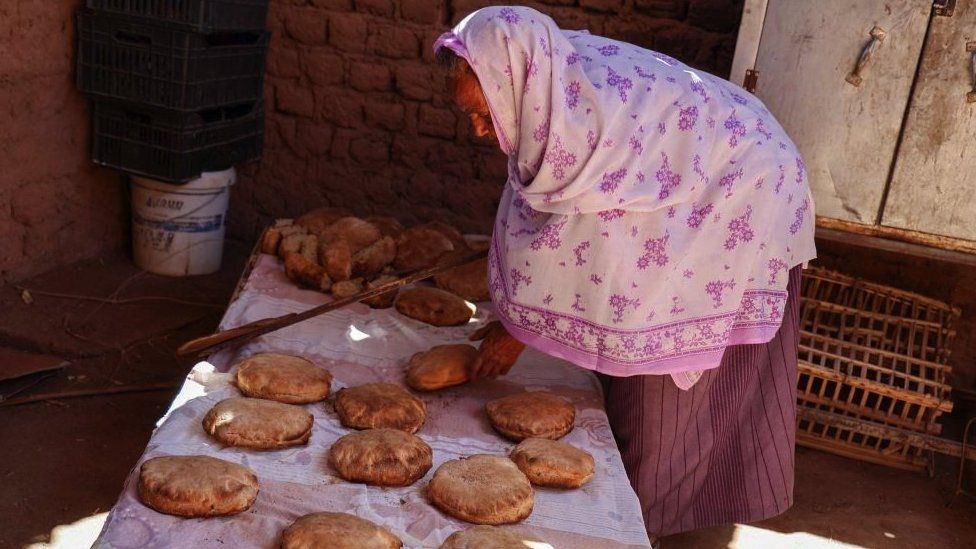 Местный житель Наамат Джабал Сайид Хасан, 75 лет, печет хлеб в глинобитной хижине, как она делает каждый день, чтобы предложить людям, спасающимся бегством из раздираемого войной Судана, проезжающим через северный город Вади Халфа
