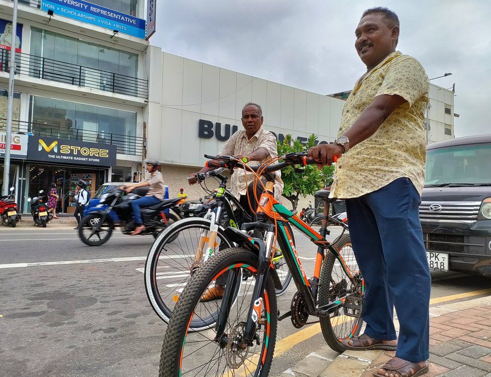 Two men hold new bicycles on the street in front of a shopping area