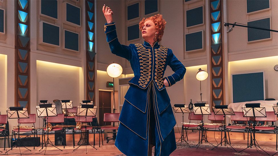 A villainous character wears a floor-length blue dress with ruffles on the lower half and elaborate gold stitching on the top that is reminiscent of Napoleon-era costume. She's standing with her back to an orchestra rehearsal room, with neat rows of chairs and sheet music stands in a v-shape formation. A boom microphone hangs from the ceiling. The character is posing side-on, their chin tilted upwards, with one hand on their hip and the other hand raised, palm up, as if encouraging applause or adulation.