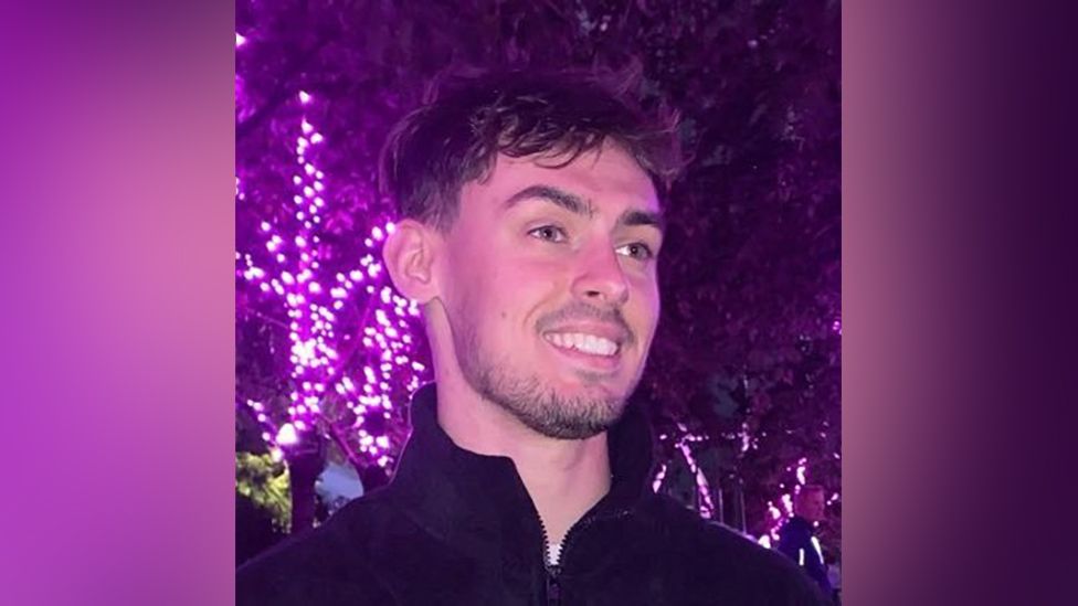 A man with short, dark tousled hair wears a high-necked zip-up fleece as he stands, smiling, in front of a tree decorated with bright purple lights.