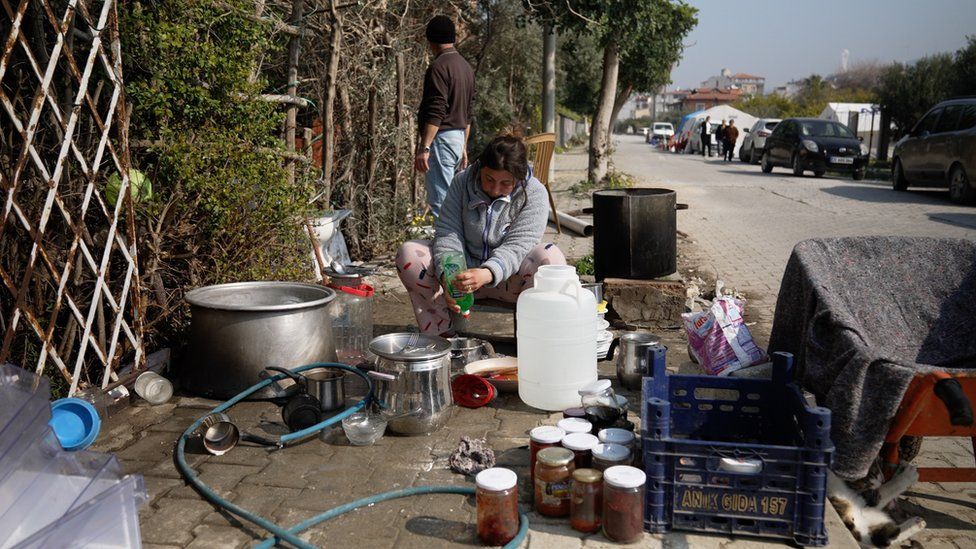 Songul Yucesoy washing dishes outside, in the shadow of her ruined home