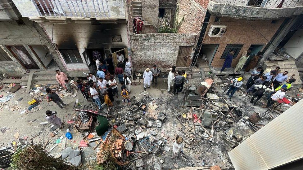 People stand in a street in Jaranwala with debris all over the ground and damaged houses in the background