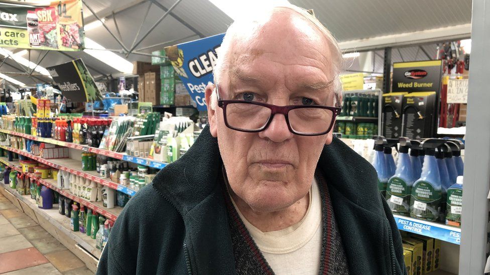 Colin Hoare looking into the camera, wearing glasses and a work fleece. Standing in an aisle of the garden centre which has liquids like weedkillers