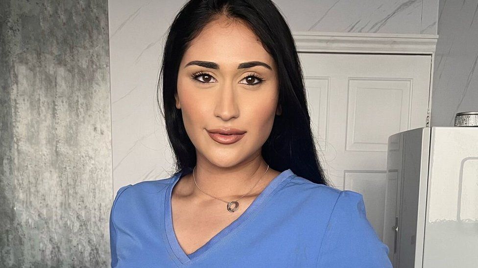 Nyrah Saleem, looking at the camera and slightly smiling, she is wearing a blue pharmacist's uniform. The background has a white door.