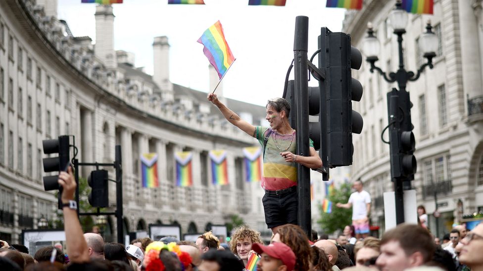 A man stands on top of a traffic light in Regent Street as thousands watch the Pride Parade