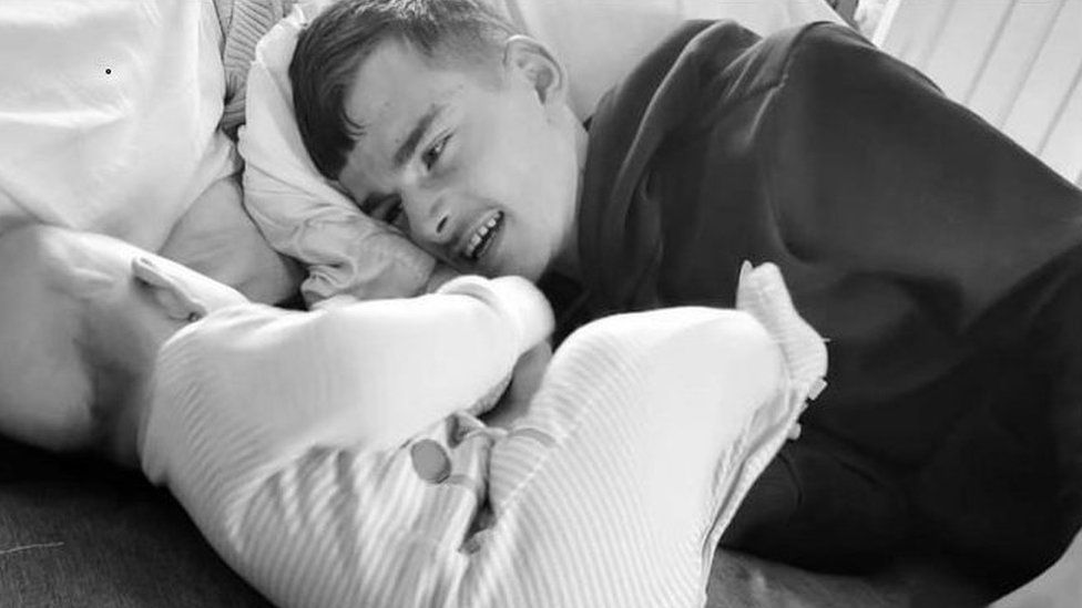 Black and white image of Connor Burden. He looks across to a baby.