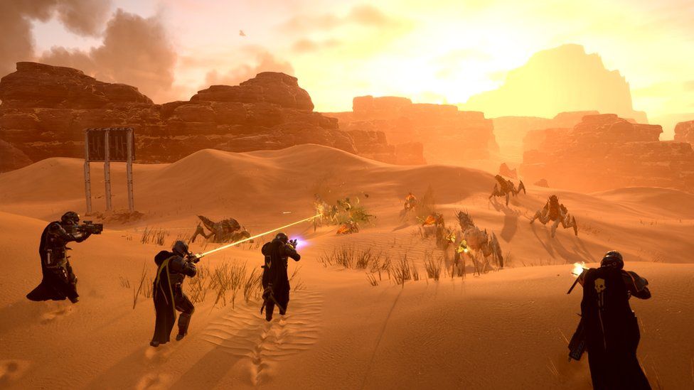Four armoured characters wielding futuristic weapons advance on a group of six insect-like creatures with spiky segmented legs and exaggerated armoured bodies in a desert landscape. The orange sunset highlights sand and dust, creating a hazy atmosphere. The corpse of one creature burns on the sand while another is pictured as it explodes on contact with a yellow laser beam emanating from the barrel of one soldier's gun.