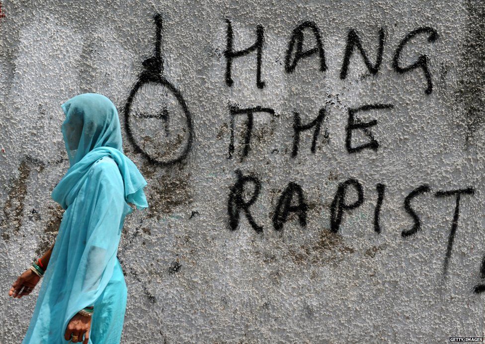 Woman stands near graffiti saying Hang the Rapist