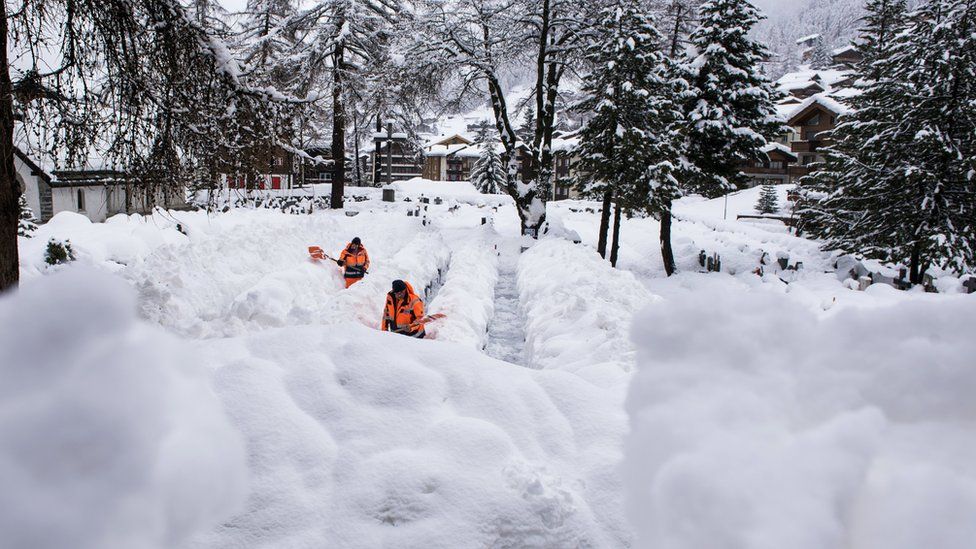 Two workers in fluorescent orange remove the snow off a path in Zermatt - it is easily up to their waist in places as they shovel it aside