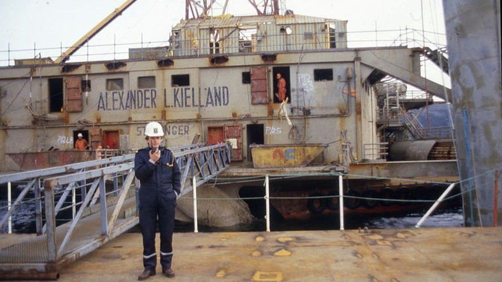 Old picture of a man in overalls and a hard hat standing in front of the wreck of the Kielland