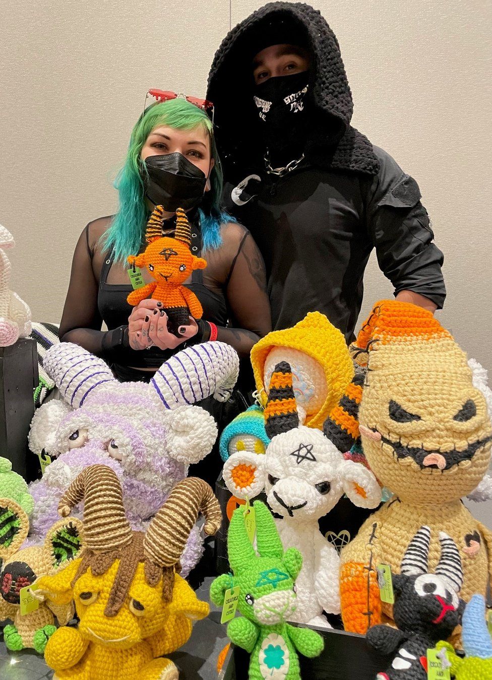 Little Nicky, who has green hair and wears black, is smiling in a Covid-19 mask. She stands in front of a table covered in crocheted toys with horns, flanked by her partner who wears lack and a Covid mask.