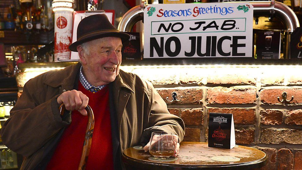 A man sitting at a bar, having a drink - a sign beside him reads: Seasons greetings - no jab, no juice