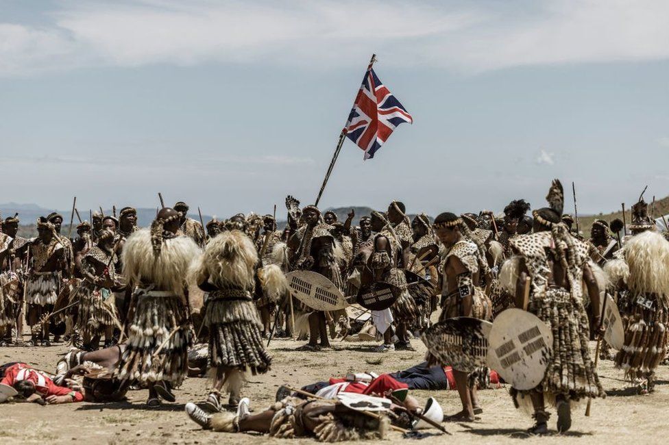 Amabutho Zulu regiments dressed in customary attire re-enact a battle. One man is holding the British flag.