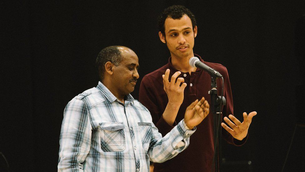 A young man of African origin standing at a microphone next to an older man who is an interpreter for him.