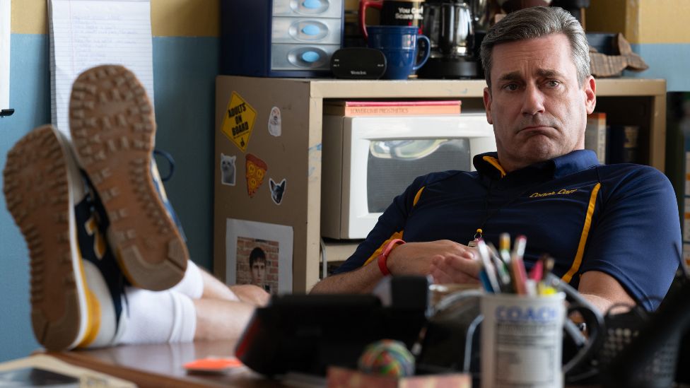 Jon Hamm as Coach Carr in 2024's Mean Girls. Coach Carr, who has greying hair and wears a blue and orange sports kit, leans back in his chair, his feet on the desk in front of him. He's pictured in his office with a stern expression.