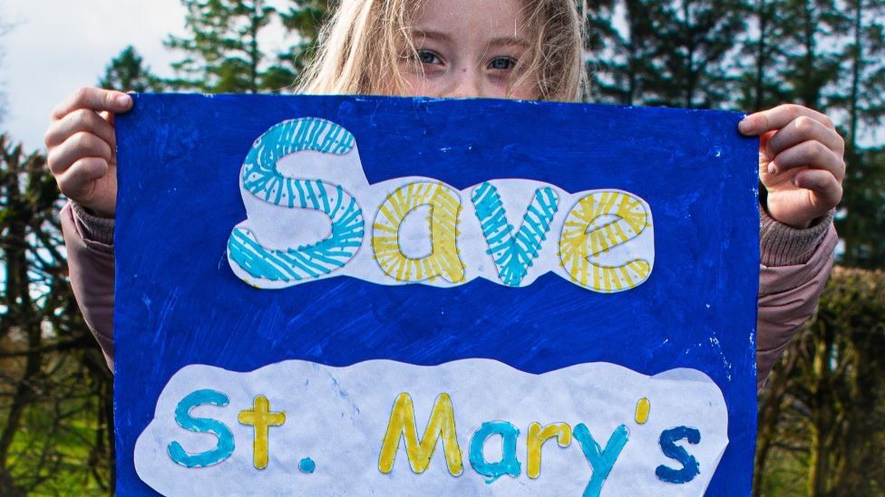A child holding a sig that reads: Save St Mary's