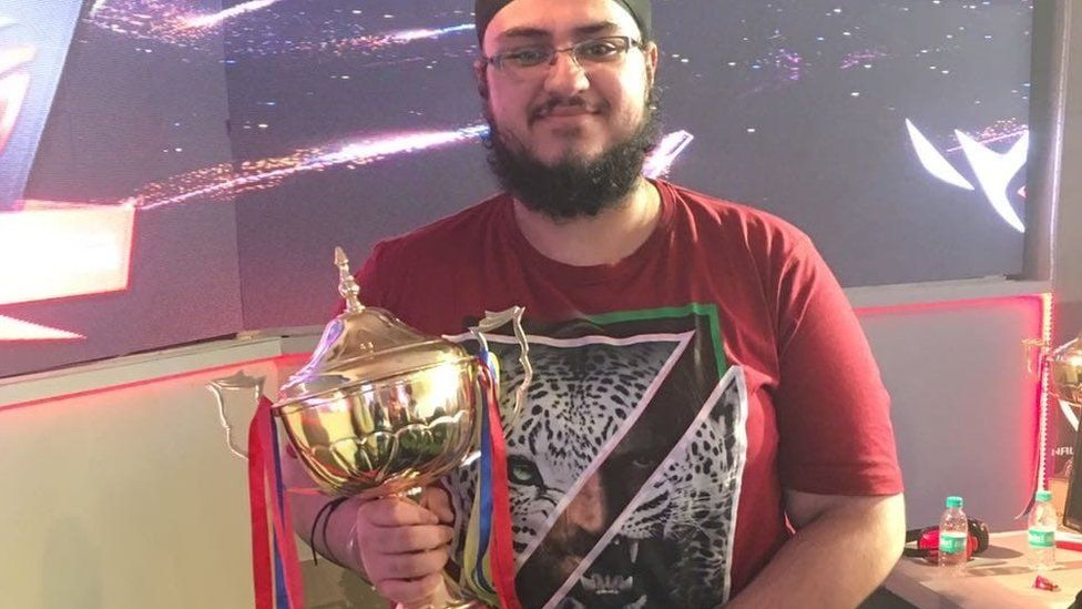 A young Asian man with a bushy beard, glasses holds a large golden trophy. He's wearing a patka - a religious head covering worn by Sikh men. It sits tight on the head and has a topknot that sits on the crown of the skull. The trophy has elaborate handles on either side and blue and white ribbons dangling off them. The man's smiling, and wears a red t-shirt with a white tiger face design on it. The picture is cut in half diagonally, and underneath the face of a male is visible.
