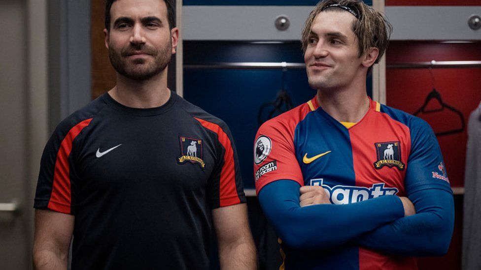 (L-R) Brett Goldstein as his character Roy Kent in the dressing room with Phil Dunster as Jamie Tartt. Roy has a football training top on and Jamie has his football kit on and is smirking at Roy